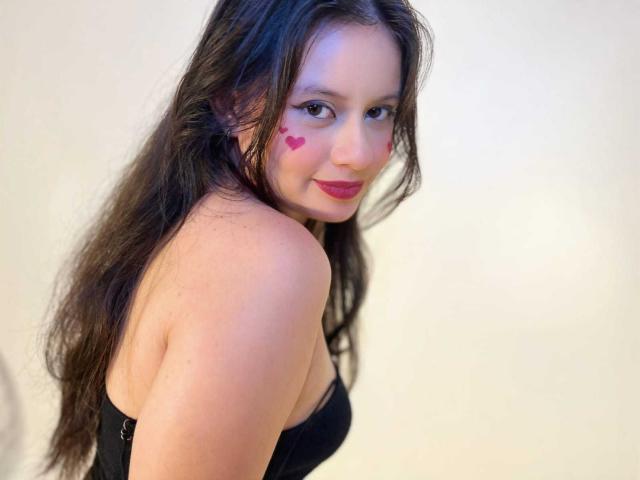 LunaRomano with long brown hair, black strapless top, pink heart makeup on cheeks, looking over shoulder, smiling, showing bare shoulders and upper back.
