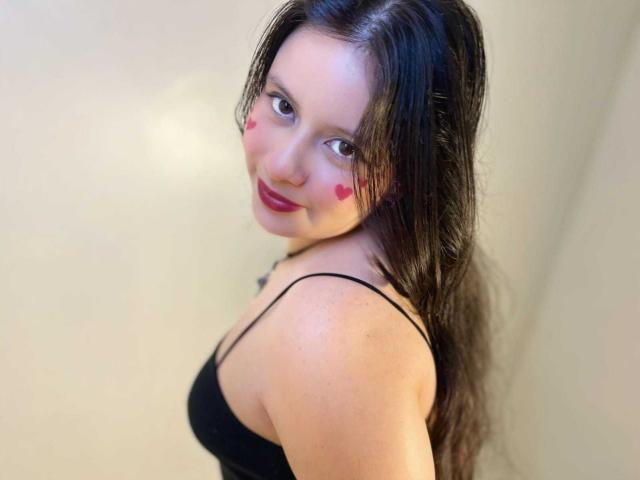 LunaRomano with wet black hair, red heart makeup on cheeks, black spaghetti strap top, smiling at camera, showing her tits. Soft light, slight upper body angle.