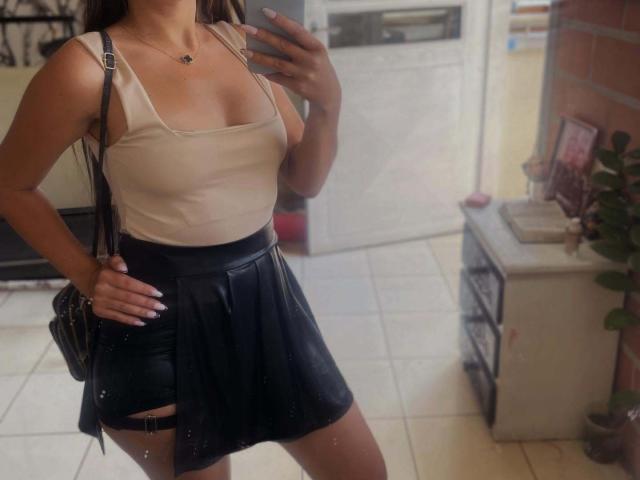 MoreForMyPussy with tan top showing boobs, black leather skirt, hand on hip, phone in hand, brown hair, necklace, black bag, standing in room with tiled floor.