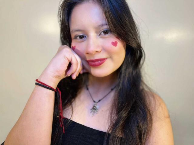 LunaRomano avec un maquillage en forme de cœur sur ses joues, un haut noir à épaules dénudées, de longs cheveux foncés, du rouge à lèvres rouge, une main près de son visage, un bracelet rouge et noir, un collier avec un pendentif..