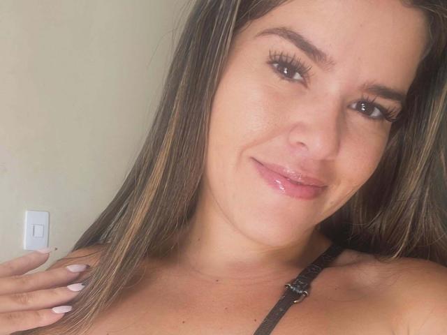 MoreForMyPussy with long brown hair, brown eyes, glossy pink lips, and white nails, wearing a black strap, smiling slightly, hand on shoulder.