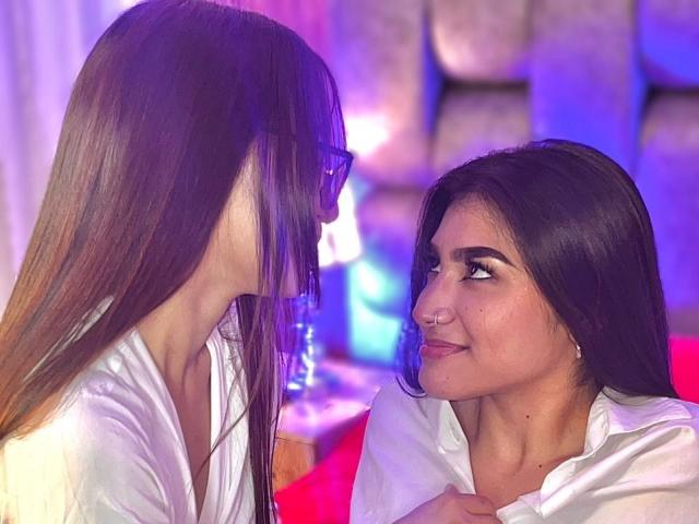 AfroditaAndHermione, con el pelo largo y castaño, se inclina, con la mirada fija, sonriendo a AfroditaAndHermione, con el pelo oscuro, camisa blanca y piercing en la nariz, que está sentada en una luz suave de color azul y rosa..