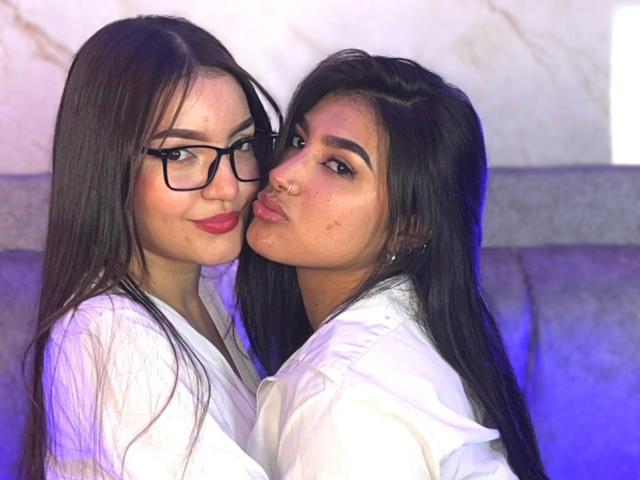 Afrodita e Hermione, con lunghi capelli neri, occhiali e camicie bianche, si baciano il naso, entrambe con rossetto rosso; Afrodita ha un piercing al naso, Hermione ha gli occhi azzurri, in un ambiente accogliente..