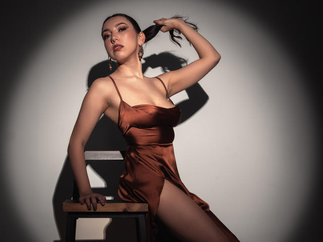 Mia Reed in een zijdezachte bruine jurk, die haar haar optilt, haar borsten laat zien, zittend op een stoel, met een schaduw op de muur, er sexy uitzien, met een spotlight op haar gericht, op een donkere achtergrond..
