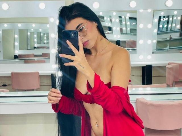 AmilyBella with long black hair, red off-shoulder top, small tits, flat stomach, and navel piercing, taking a mirror selfie in a lit room with round mirrors.