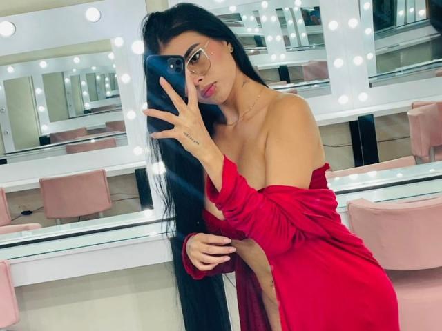 AmilyBella with long black hair, glasses, and red dress, holding phone, showing bare shoulder and side of boob, mirror with lights in background, tattoo on hand.