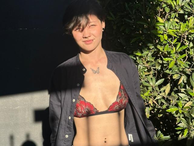 LissyLuxe with short black hair, black open shirt, and red lace bra, showing small tits and a butterfly tattoo above her chest, standing in sunlight with green leaves.