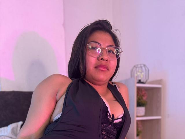 SsolSaenz a la peau marron clair, les cheveux noirs, porte des lunettes et est vêtue d'un haut noir sans manches avec un soutien-gorge en dentelle qui laisse entrevoir de petites seins. Elle est assise sous une lumière légèrement rosée..
