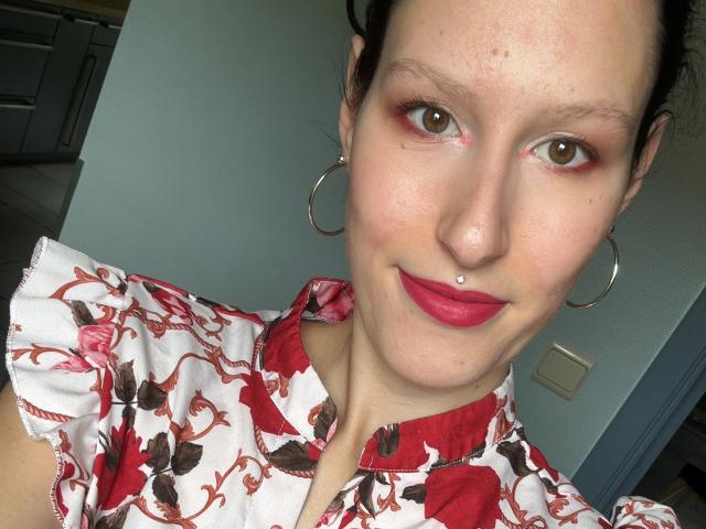JadeFrenchKiss, avec rouge à lèvres rouge, nez percé, boucles d'oreilles cerceaux et chemise à fleurs, a les yeux verts et une peau légèrement constellée de taches de rousseur, et sourit à l'appareil photo..