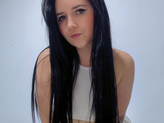 Kkatalina with long black hair, white crop top, and subtle makeup, looking down with a slight pout, showing her bare shoulders and a hint of her tits.