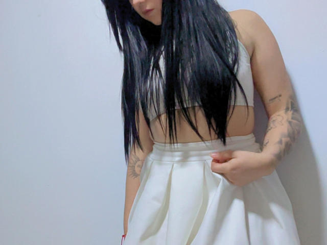 Kkatalina, con lunghi capelli neri, un top bianco e una gonna bianca, un tatuaggio sul braccio destro, tiene la gonna, mostrando un po' delle sue mutandine bianche, in piedi di fronte a una parete chiara..