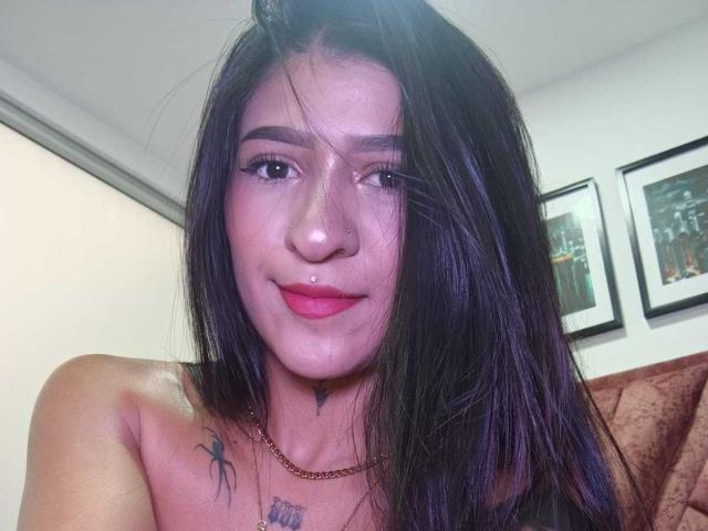 LuciadelaVega with dark purple hair, pink lipstick, and a small drop of white fluid on her chin, smiling, showing a spider tattoo on her left chest.