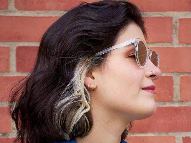 NyxVale has dark wavy hair with white highlights, wears round clear glasses, nose ring, and pink lipstick, looking side profile, smiling slightly, with a red brick background.