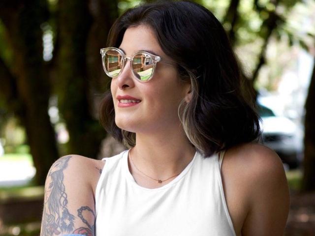 NyxVale, usando óculos de sol espelhados, uma camiseta branca e com o braço tatuado, sorrindo ao ar livre. Ela tem cabelo escuro e ondulado, um rosto bonito e um pequeno colar de ouro..