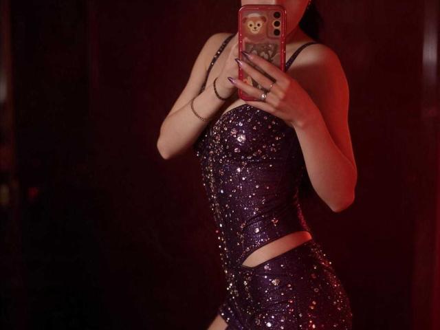 Boorbuja wearing a sparkly purple dress with sequins, showing a cutout at the waist, holding a phone with a teddy bear case, reflecting in a mirror.