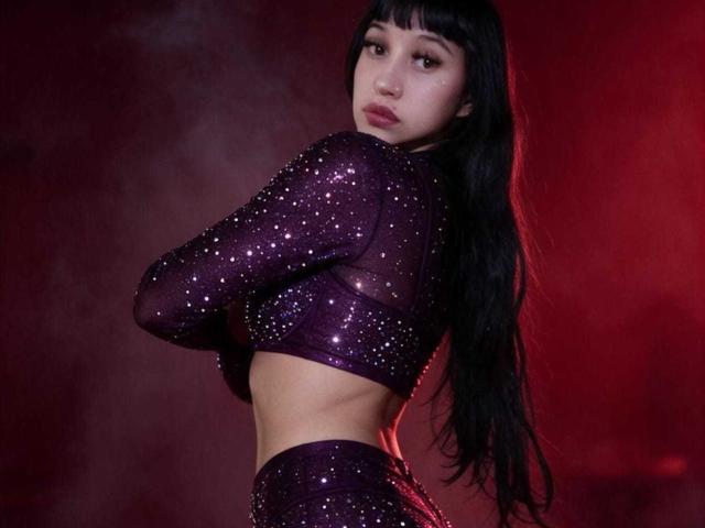 Boorbuja with long black hair, wearing a purple sparkly crop top and matching pants, arms crossed, showing a slim waist and small tits, red-lit background.