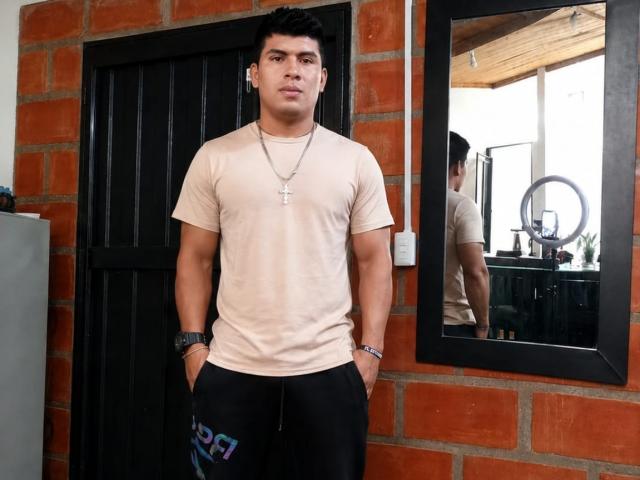 KairoXL, com uma estrutura física forte, vestindo uma camiseta bege, calças pretas, com as mãos nos bolsos, um colar, um relógio e cabelo preto curto, está em frente a um espelho..