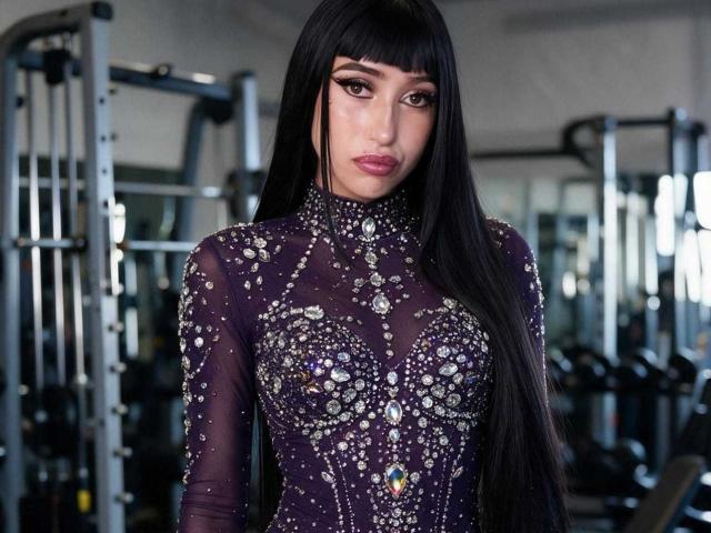 Boorbuja, con lunghi capelli neri, indossa un abito viola trasparente con intricati motivi in argento e pietre preziose, che copre il seno e i glutei. Sullo sfondo, si vede un ambiente da palestra..