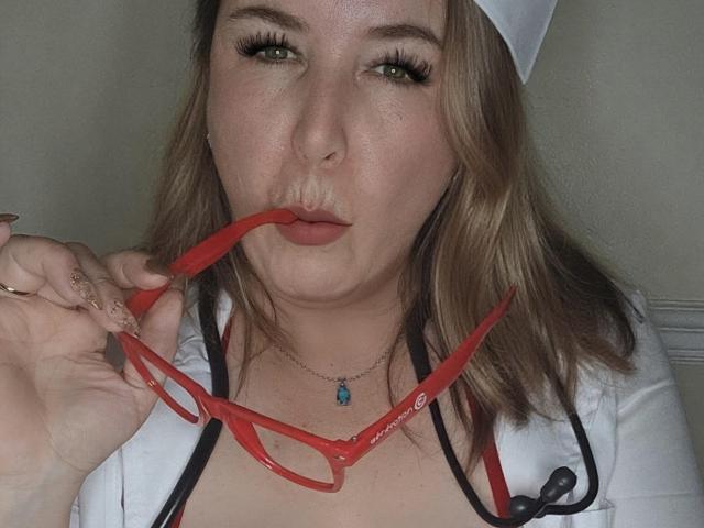 TitanicTits with light brown hair, wearing a white nurse cap and shirt, puffs out red glasses with her lips, showing off her nose ring and long nails.