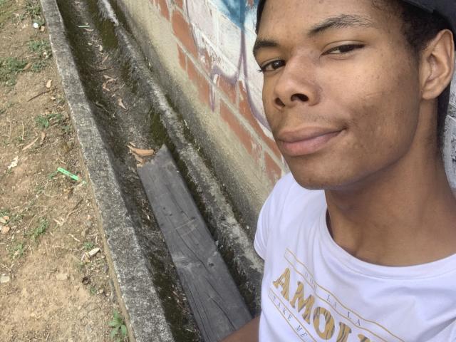 AndyCris has a medium brown skin tone, short curly hair, a slight smirk, and is wearing a white t-shirt with a gold design. He's standing next to a dirty wall.