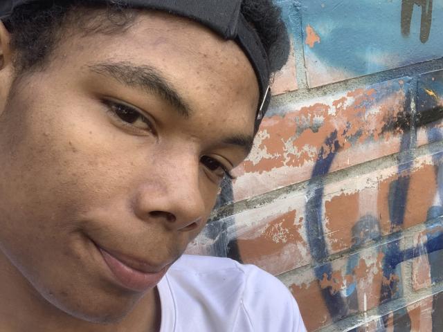 AndyCris has a small smile, brown eyes, and short black hair with a black cap, wearing a white shirt, against a graffiti-covered brick wall.