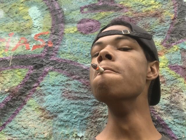 AndyCris, dark-skinned, wearing a backward black cap, has a small red and black cigarette in his mouth, looking up with a slight smirk, against a graffiti wall.