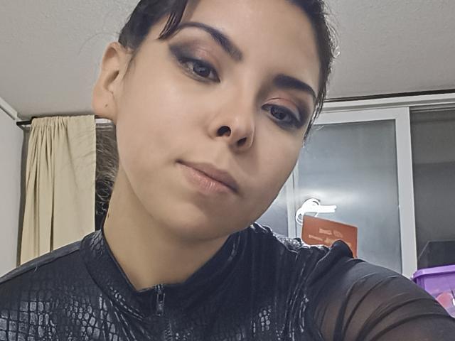 IviHetairaa, con sopracciglia scure, eyeliner e un sorriso appena accennato, indossa una maglia nera, lucida e con una texture particolare, e ha i capelli scuri raccolti in un'acconciatura morbida, mentre guarda verso la fotocamera..