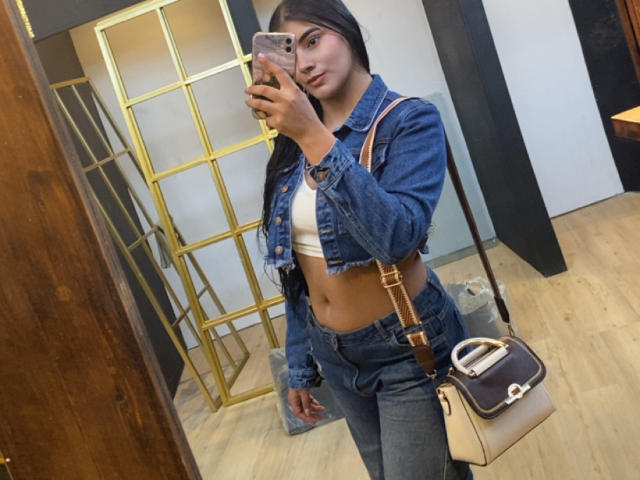 AlaraKey with long black hair, denim jacket, white crop top, gray jeans, holding phone, beige bag, mirror selfie, medium tits, slim, light brown skin, wooden floor, gold divider.