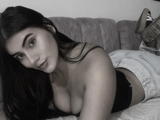 Black-and-white photo of AlaraKey lying on a couch, wearing denim shorts and a black top, showing her big boobs, with long dark hair and a slightly curious expression.