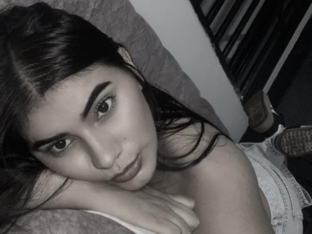 AlaraKey has dark, straight hair and is lying on a bed, looking at the camera with a slight, sultry smile. Her hand rests on her shoulder, showing her tits.