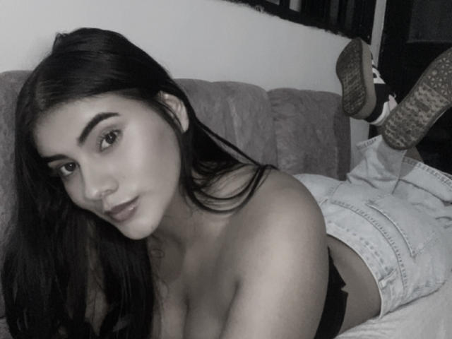 AlaraKey with long dark hair, lying on a couch, topless, showing her tits, wearing jeans and socks, legs up, looking at the camera, in a grayscale photo.
