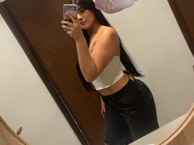 AlaraKey, con cabello negro largo, un top blanco y jeans negros, sosteniendo un teléfono para un selfie frente al espejo, mostrando sus pechos y glúteos, de pie en el baño..