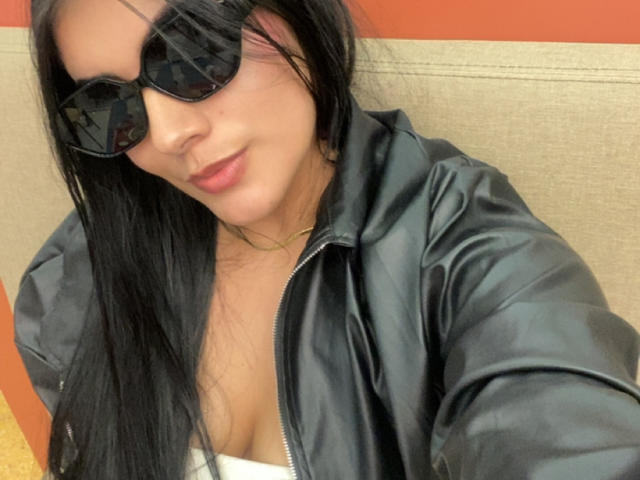 Alara, con el pelo largo y negro, usando gafas de sol negras, una chaqueta de cuero negra y una blusa blanca, mostrando un poco de sus pechos, sonriendo sutilmente..