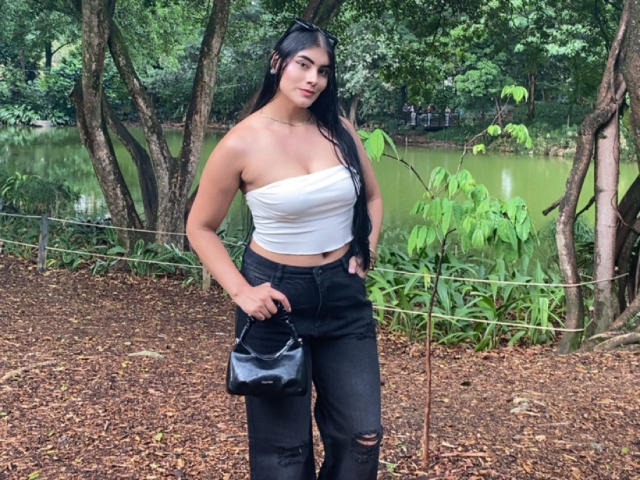 AlaraKey with long black hair, white strapless top, black ripped jeans, holding black leather purse, standing outdoors, forest background, green water, trees, brown leaf-covered ground.