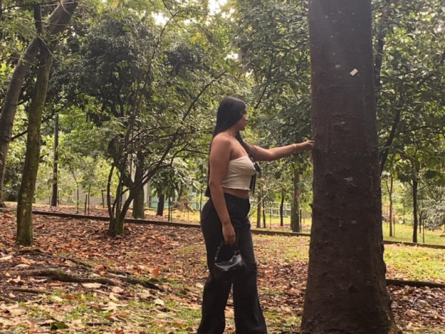 AlaraKey, con cabello negro, piel morena, vestida con un top blanco sin tirantes y pantalones negros, toca un árbol, sosteniendo un bolso negro, y está de pie en un área boscosa con hojas caídas..