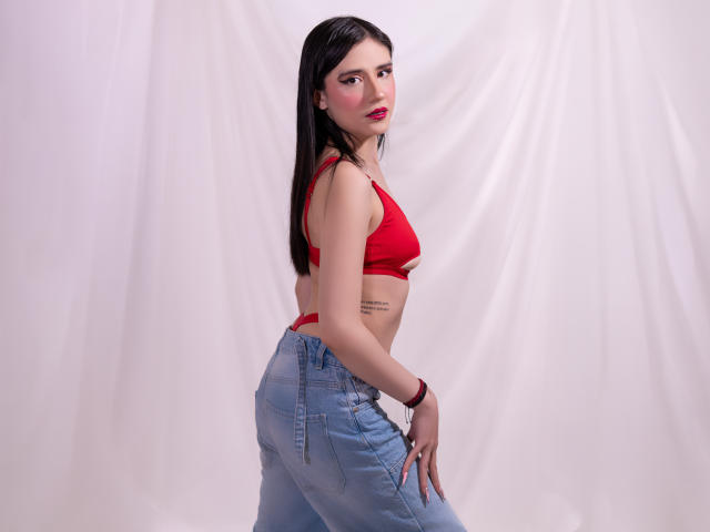 Valerie Banks met lang zwart haar, een rode bh en blauwe jeans, kijkt over haar schouder, toont kleine borsten, een subtiele tatoeage aan de zijkant, roze lippen, witte achtergrond..
