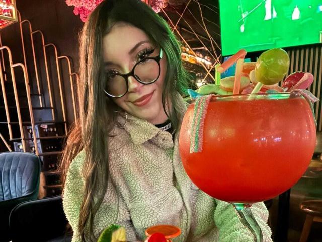 LunaValu mit Brille, grünen Augen und langen braunen Haaren, hält einen riesigen roten Cocktail mit Früchten, trägt eine flauschige beige Jacke und lächelt in die Kamera..