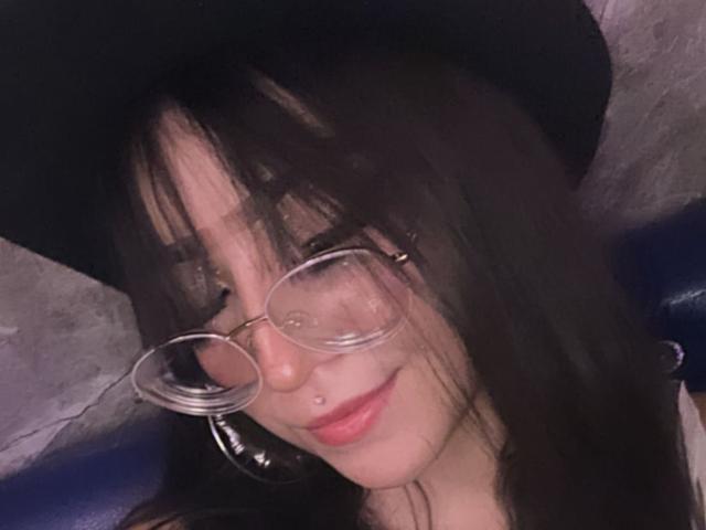 LunaValu with dark hair, wearing a black hat and round glasses, has a nose piercing and a subtle smile, showing her pink lips and slightly parted mouth.