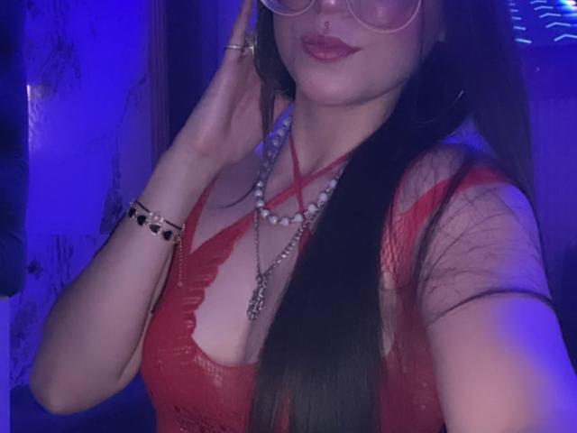LunaValu com cabelo preto longo, blusa de renda vermelha e colar, usando óculos, mão perto do rosto, iluminação azul, mostrando seus seios, batom vermelho e pulseira de pérolas..