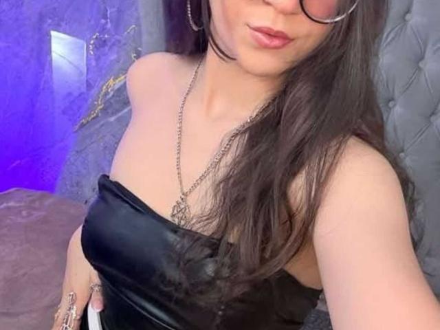 LunaValu, con cabello castaño largo, vistiendo un top negro sin tirantes, un collar de plata y gafas, haciendo pucheros con labios rosados, mostrando sus hombros desnudos y un atisbo de sus senos..