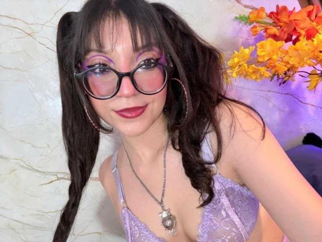 LunaValu com maria-chiquinhas pretas, óculos, sombra roxa e batom, vestindo um sutiã de renda lilás, sorrindo, brincos de argola, colar e flores vermelho-laranja ao fundo..
