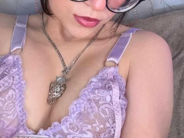 LunaValu com óculos, batom rosa, usando um sutiã de renda lilás, mostrando os seios, um colar com um pingente em formato de coração e uma corrente de prata, em um selfie casual..