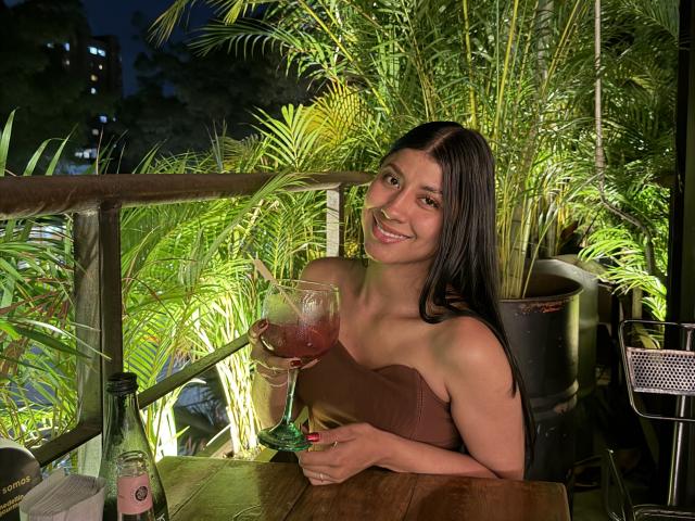MadinssonGrey with long black hair, smiling, holding a pink cocktail in a large glass, wearing a brown strapless top, sitting at a wooden table with a green bottle.
