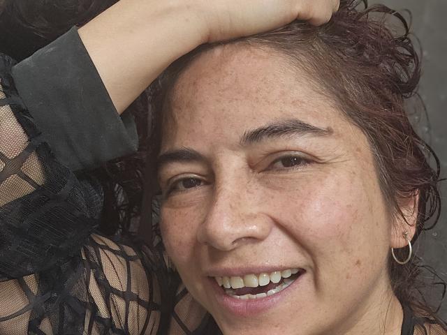 AlessiaRed with curly dark brown hair, wearing a black sheer long-sleeve top, smiling with hand on head, showing freckles, small hoop earrings, and white teeth.