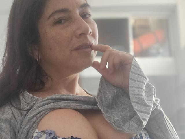 Alessia, cabelos escuros, sardas, camisa cinza, mordendo o dedo, exibindo seios grandes e nus com um sutiã azul estampado, levemente levantados. Luz suave, expressão confiante..