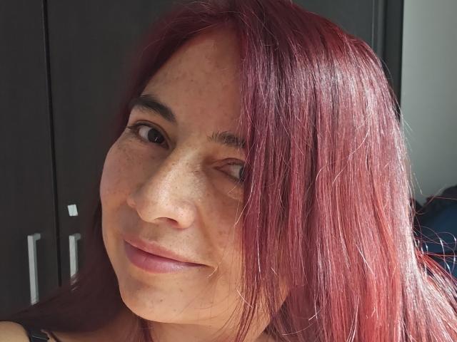 AlessiaRed with red hair, winking, smiling, freckled face, brown eyes, and pink lips. She's wearing a sleeveless top, showing a tattoo on her left shoulder.