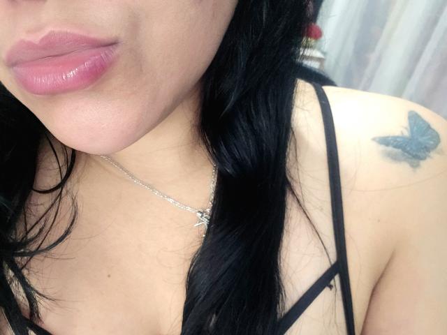 VivianaThompson's face, showing pink glossy lips, black hair, and a silver necklace with a cross. She has a blue butterfly tattoo on her right shoulder. Boobs.