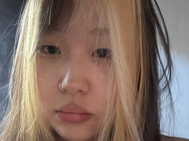 JungMcgranor has pale skin, blonde and black hair, a small nose ring, and pinkish lips. She looks bored with a slight pout. Her eyes are half-open.