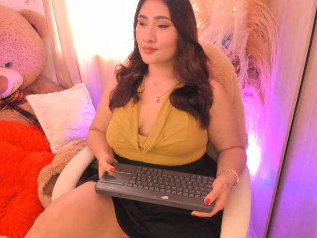 ElyRoss with big tits, wearing a yellow top and black skirt, sitting, holding a keyboard, with brown wavy hair, and a soft smile, in a cozy room.