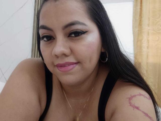 Une femme pulpeuse et voluptueuse, aux cheveux foncés, avec un eye-liner noir, un rouge à lèvres rose, de petites boucles d'oreilles, un collier en or, un débardeur noir, et un tatouage de rose sur l'épaule droite, souriant à l'appareil photo..